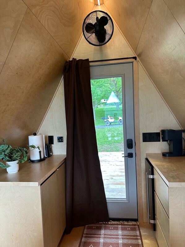 A small, triangular room with a ceiling fan, a door with a brown curtain, a small table with a plant, a coffee machine, and a view of a house and lawn outside. - The Bellefonte Campground (Bellefonte)