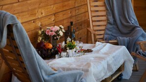 Un chalet en bois chaleureux, avec une table à manger dressée pour deux personnes, agrémentée d’une nappe en dentelle blanche, d’une bouteille de vin, d’un vase de fleurs et d’une bougie.