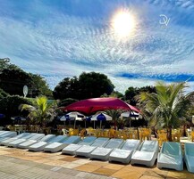 Uma área à beira da piscina com espreguiçadeiras e guarda-sóis, cercada por palmeiras e um céu limpo.