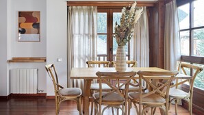 Een eetgedeelte met een houten tafel en stoelen, een vaas met gedroogde planten en een raam met gordijnen.