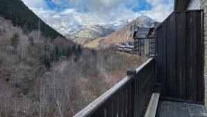 A mountain view from a balcony with a wooden railing, overlooking a valley with snow-capped mountains and a building with multiple balconies. - Casa Val de Ruda Luxe 71 by FeelFree Rentals (Baqueira)