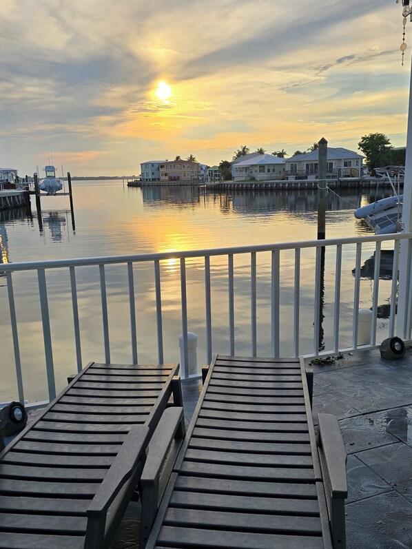 A waterfront view with a wooden deck, a boat docked on the water, and buildings in the background. - Beach Waterfront Home with Private Beach Access & Boat Ramp/Dock in Paradise! (Jensen Beach)