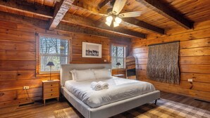 Ein Schlafzimmer einer Holzhütte mit Bett, Nachttischen, Deckenventilator und einem Fenster mit Jalousien.