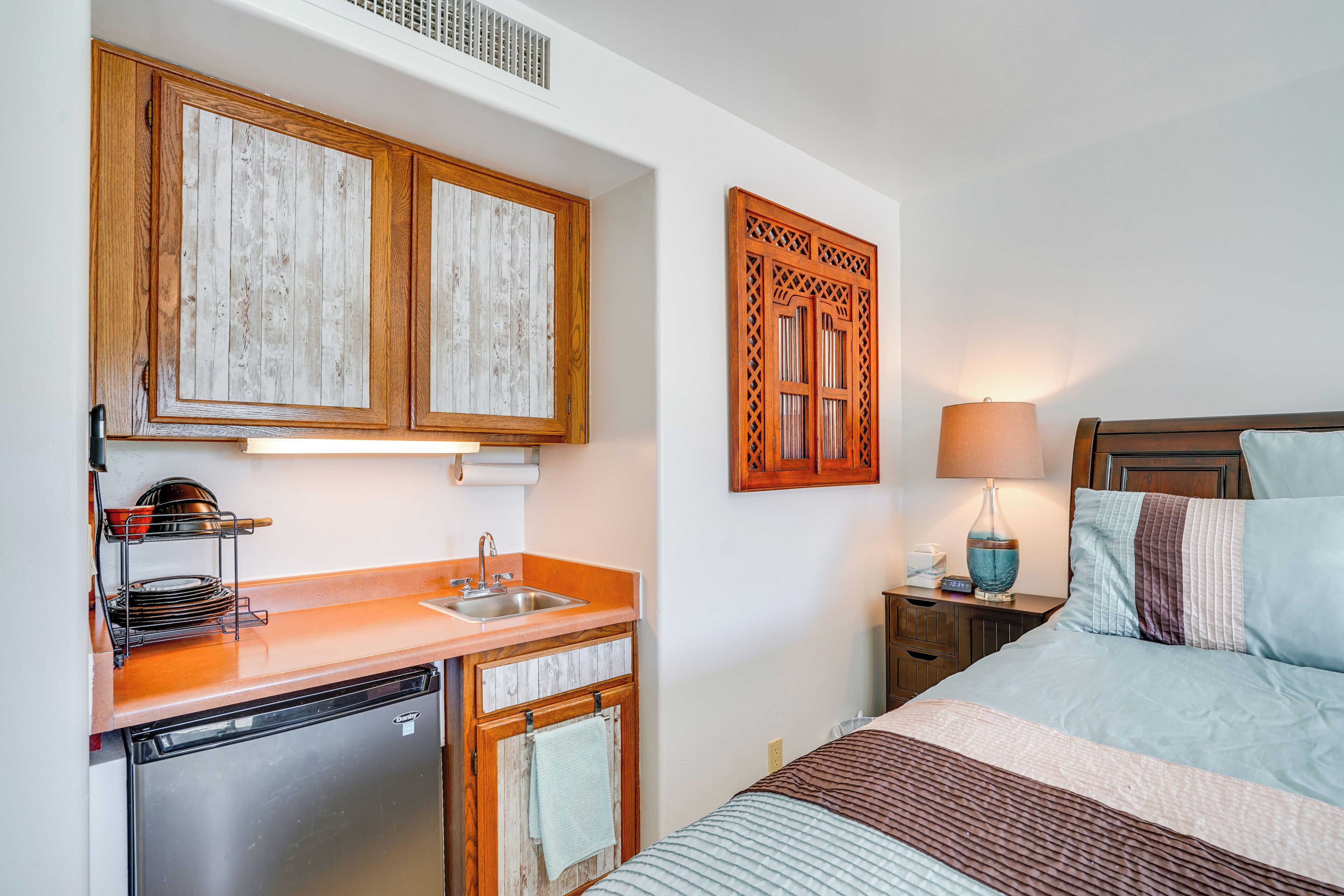 Ein kompaktes Hotelzimmer mit Küchenzeile, einem Bett mit blau-weiß gestreifter Decke, einem Holz-Kopfteil und einem Nachttisch mit Lampe.