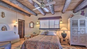 Een slaapkamer met een houten plafond, een bed met beddengoed met een patroon, een nachtkastje met een lamp, een dressoir met decoratieve deuren, en een raam met zonwering.
