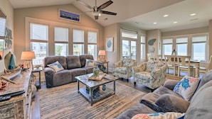 A spacious living room with a ceiling fan, large windows, a fireplace, and a dining area with chairs and a table. - Charming Surf City Home w/ Elevator: Walk to Beach (Holly Ridge)
