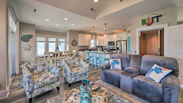 A spacious living area with two patterned armchairs, a gray sofa, and a coffee table with decorative items. A kitchen with white cabinets and a dining area with a wooden table and chairs are visible in the background. - Charming Surf City Home w/ Elevator: Walk to Beach (Holly Ridge)