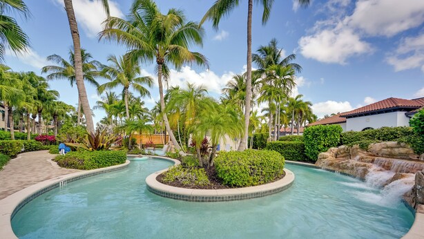 A swimming pool with a waterfall feature, surrounded by palm trees and lush greenery. - Luxe Naples Bay Resort Condo: Near Fifth Ave! (Naples)