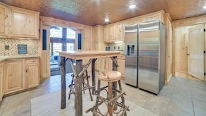 Uma cozinha com armários de madeira, uma ilha central com banquetas, uma geladeira de aço inoxidável e um teto decorativo.