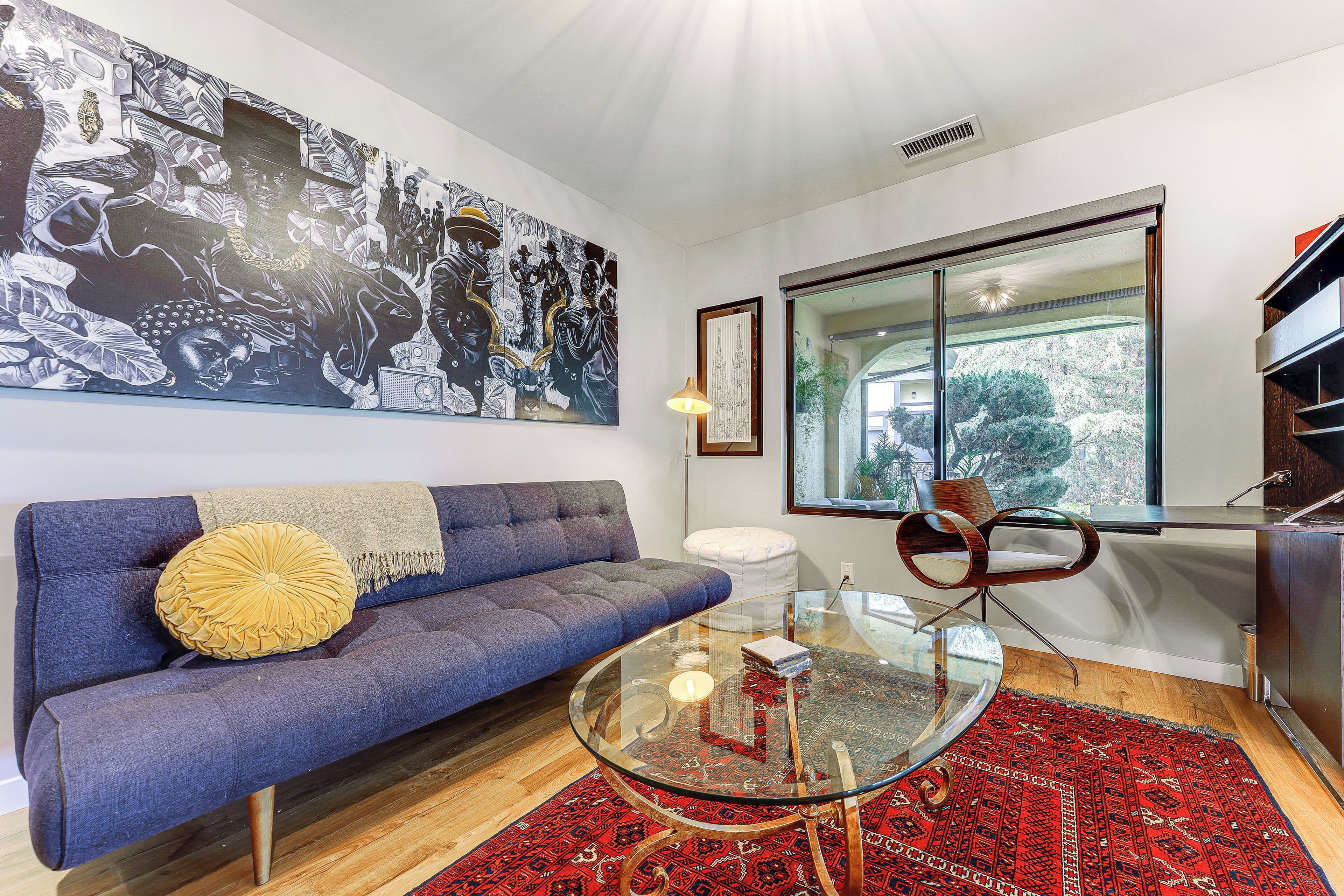 Een moderne woonkamer met een blauwe bank, een glazen salontafel, een rood tapijt met een patroon en een groot zwart-wit kunstwerk aan de muur.