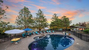 A swimming pool with lounge chairs and umbrellas, surrounded by trees and a sunset sky.