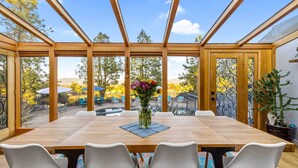 A dining area with a wooden table and white chairs, a vase of flowers, and a view of the outdoors through large windows.