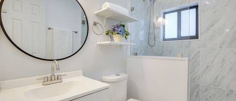 Un baño con espejo redondo, lavamanos blanco, inodoro y ducha. Hay estantes con toallas y una planta en maceta.