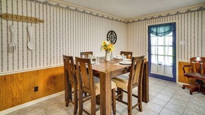 Une salle à manger avec une table et des chaises en bois, un vase rempli de fleurs et un panneau décoratif représentant une cuisine accroché au mur.