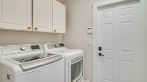 A laundry room with two white washing machines and a dryer, a white door, and a thermostat on the wall. - Florida Getaway w/ Heated Pool, Bar & Fire Table! (Cape Coral)