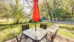 Un patio exterior con una mesa de vidrio y sillas de metal, rodeado por una cerca de malla y árboles.