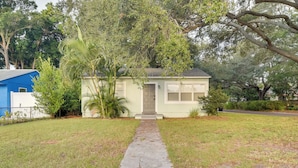 A small, single-story house with a front door, a narrow stone pathway leading to it, and a lawn with scattered trees and shrubs. - Pet-friendly Gulfport Home: Walkable Location! (St. Petersburg)