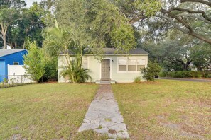 A small, single-story house with a front door, a narrow stone pathway leading to it, and a lawn with scattered trees and shrubs. - Pet-friendly Gulfport Home: Walkable Location! (St. Petersburg)