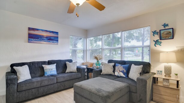 A living room with a gray sectional sofa, a wooden coffee table, and a ceiling fan. There are large windows with blinds, a painting of a sunset, and a small table with a lamp and decorative items. - Pet-friendly Gulfport Home: Walkable Location! (St. Petersburg)