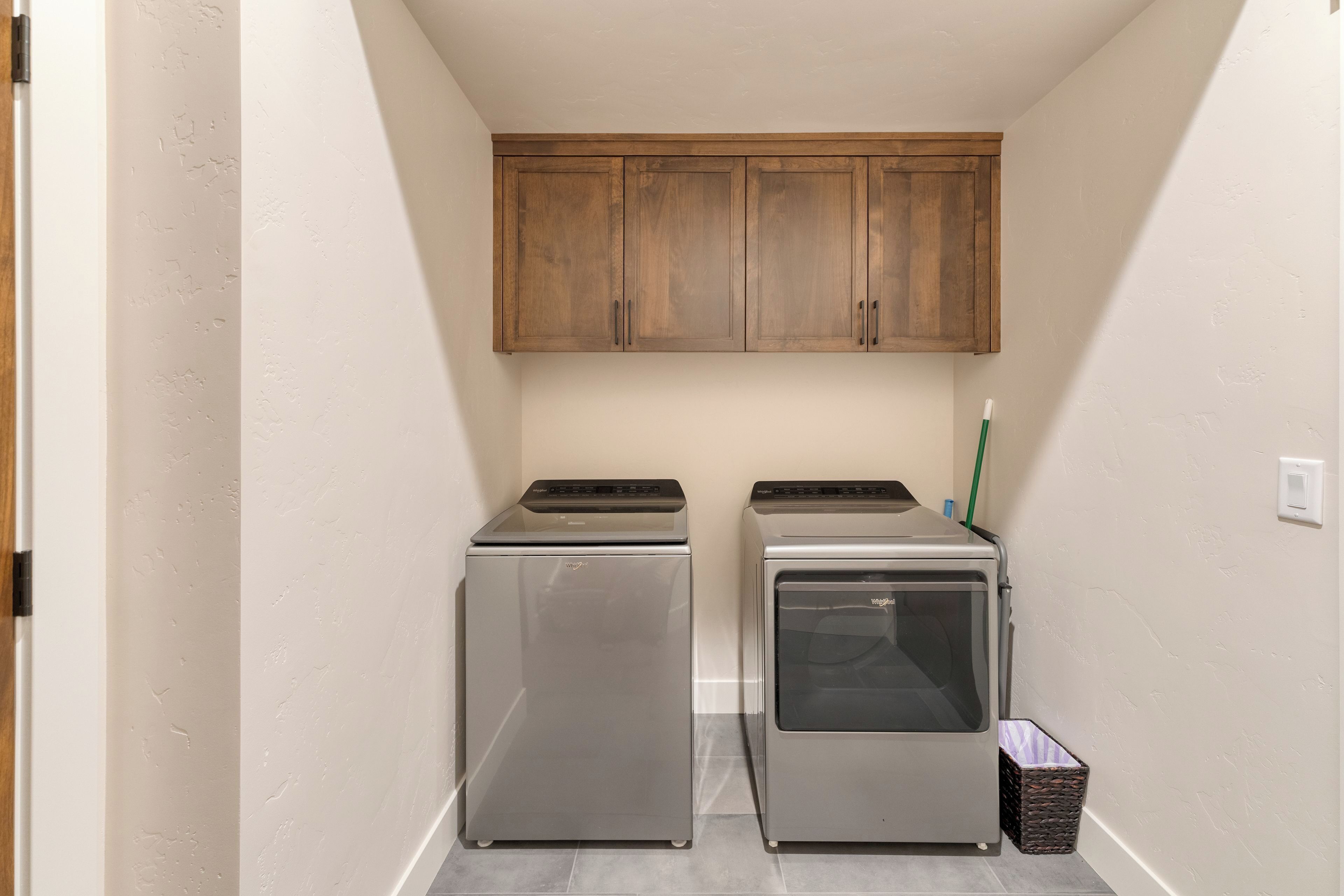 A laundry room with two stainless steel appliances, wooden cabinets above, and a basket on the floor.
