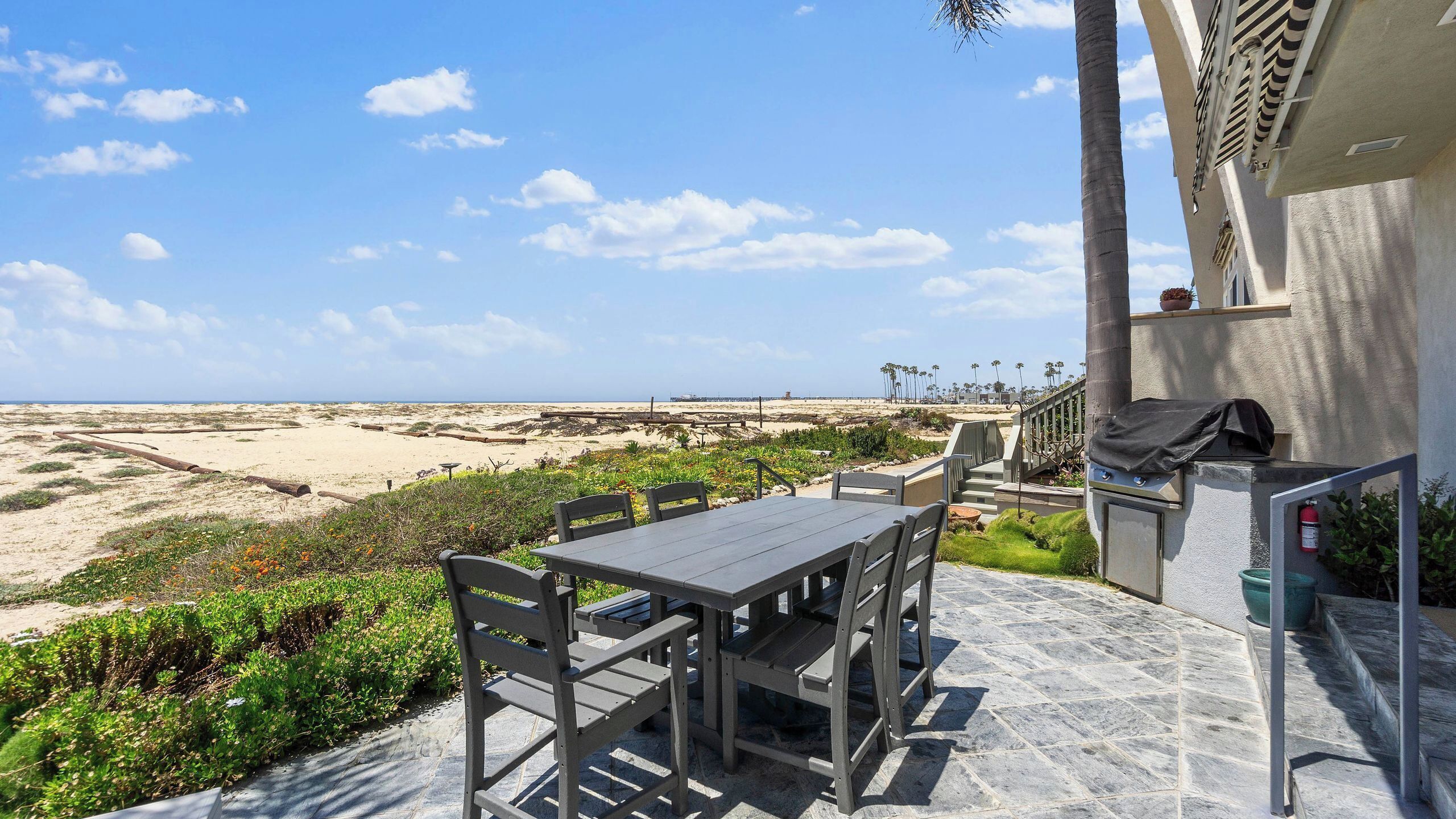 A patio with a table and chairs, overlooking a beach with dunes and palm trees.