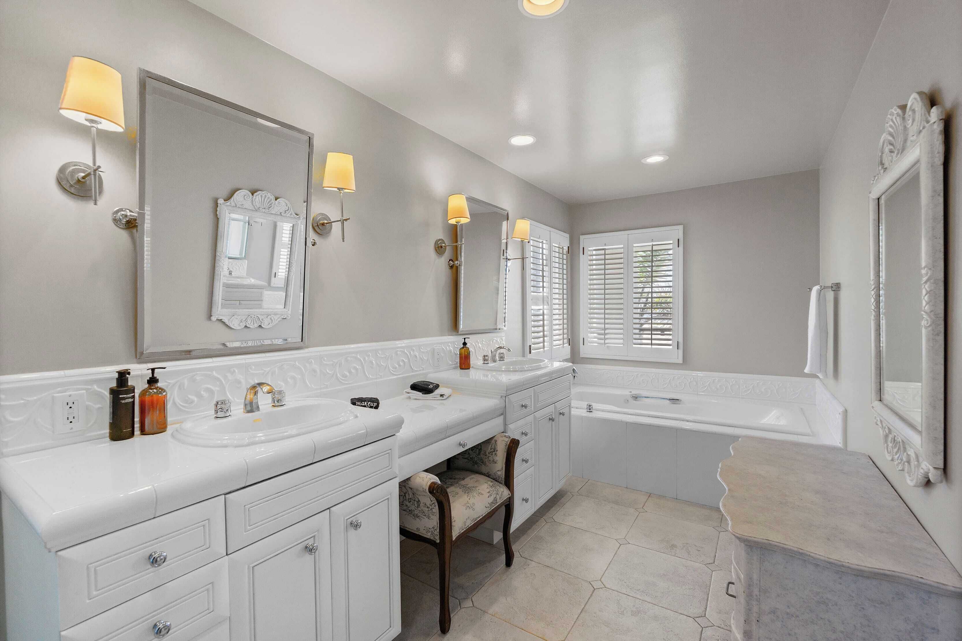 A bathroom with two sinks, a large mirror, and a bathtub. There are wall sconces and a window with blinds.