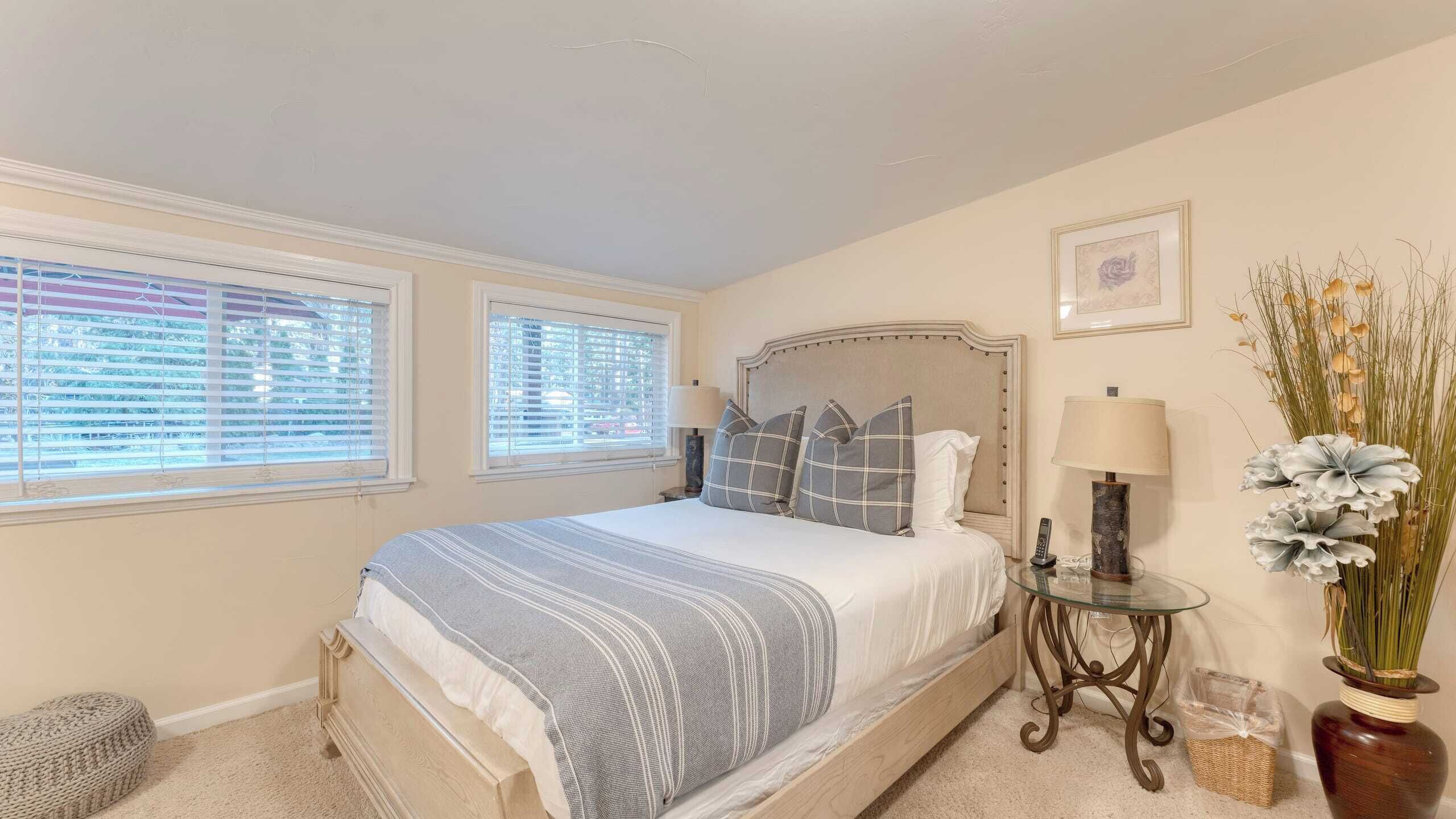 A bedroom with a bed, two windows, a bedside table, a lamp, a vase with flowers, and a framed picture on the wall.