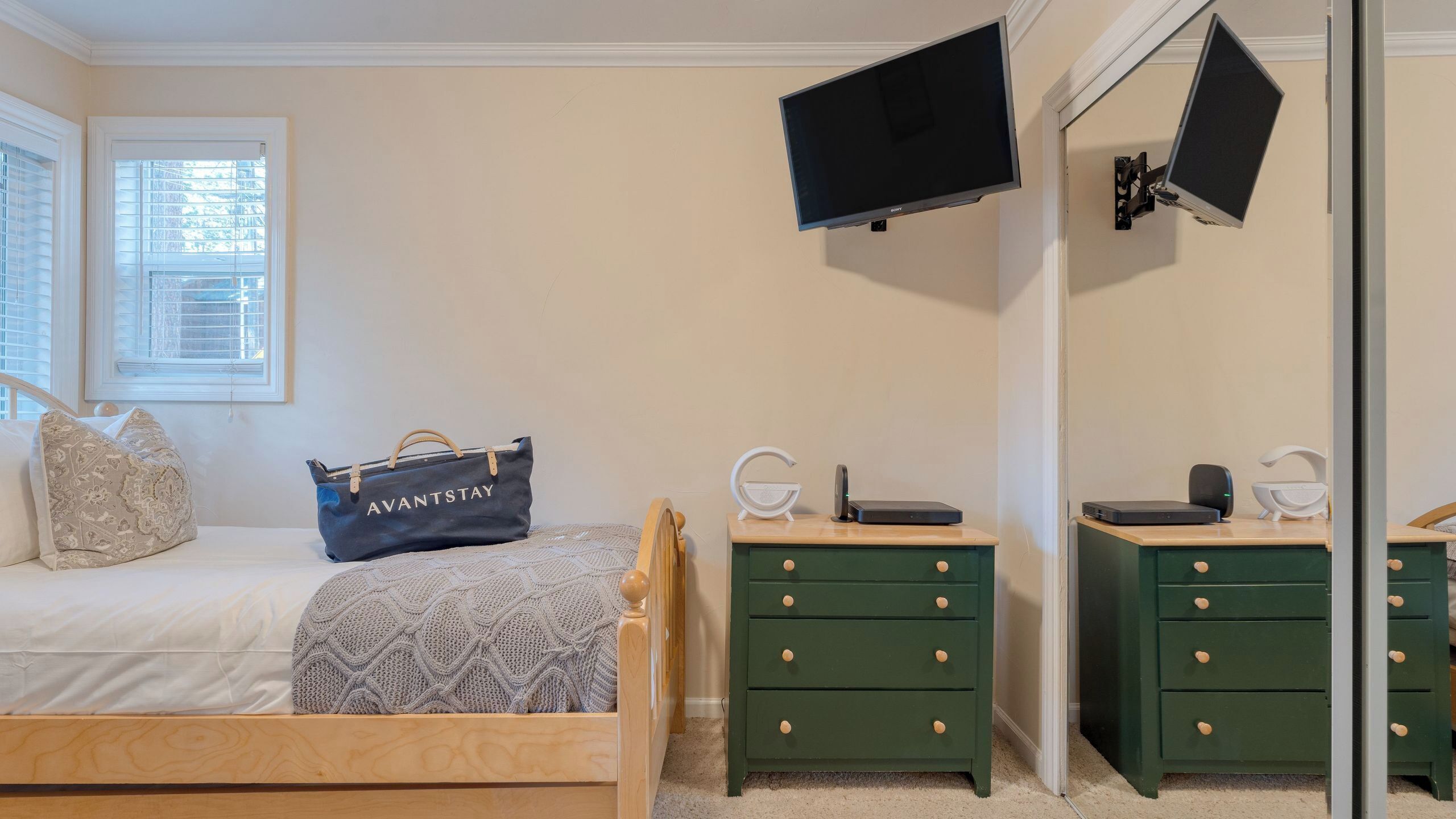 A bedroom with a bed, a bedside table, a dresser, a TV mounted on the wall, and a bag labeled 'AVANTSTAY'.