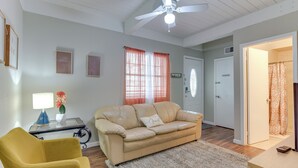 Uma sala de estar com um sofá bege, uma poltrona amarela, uma mesa de centro de vidro e um abajur, e um ventilador de teto.
