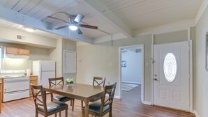 Uma sala de jantar com mesa e cadeiras de madeira, um ventilador de teto e uma cozinha ao fundo.