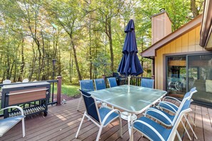 A deck with a glass table and chairs, a barbecue grill, and a covered umbrella. - Pocono Pines Gem w/ Fire Pit & On-site Creek! (Pocono Pines)