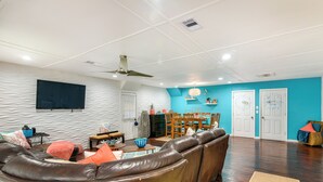 Ein Wohnzimmer mit braunem Ledersofa, einem wandmontierten Flachbildfernseher, einem Deckenventilator und einem Essbereich mit Holztisch und Stühlen.