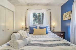 A bedroom with a bed, two bedside tables, a window with curtains, a painting on the wall, and a door.