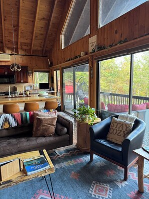Ein gemütliches Blockhaus-Interieur mit Holzbalkendecke, Essbereich, einer Couch, einem Sessel und einem großen Fenster mit Blick auf Bäume.