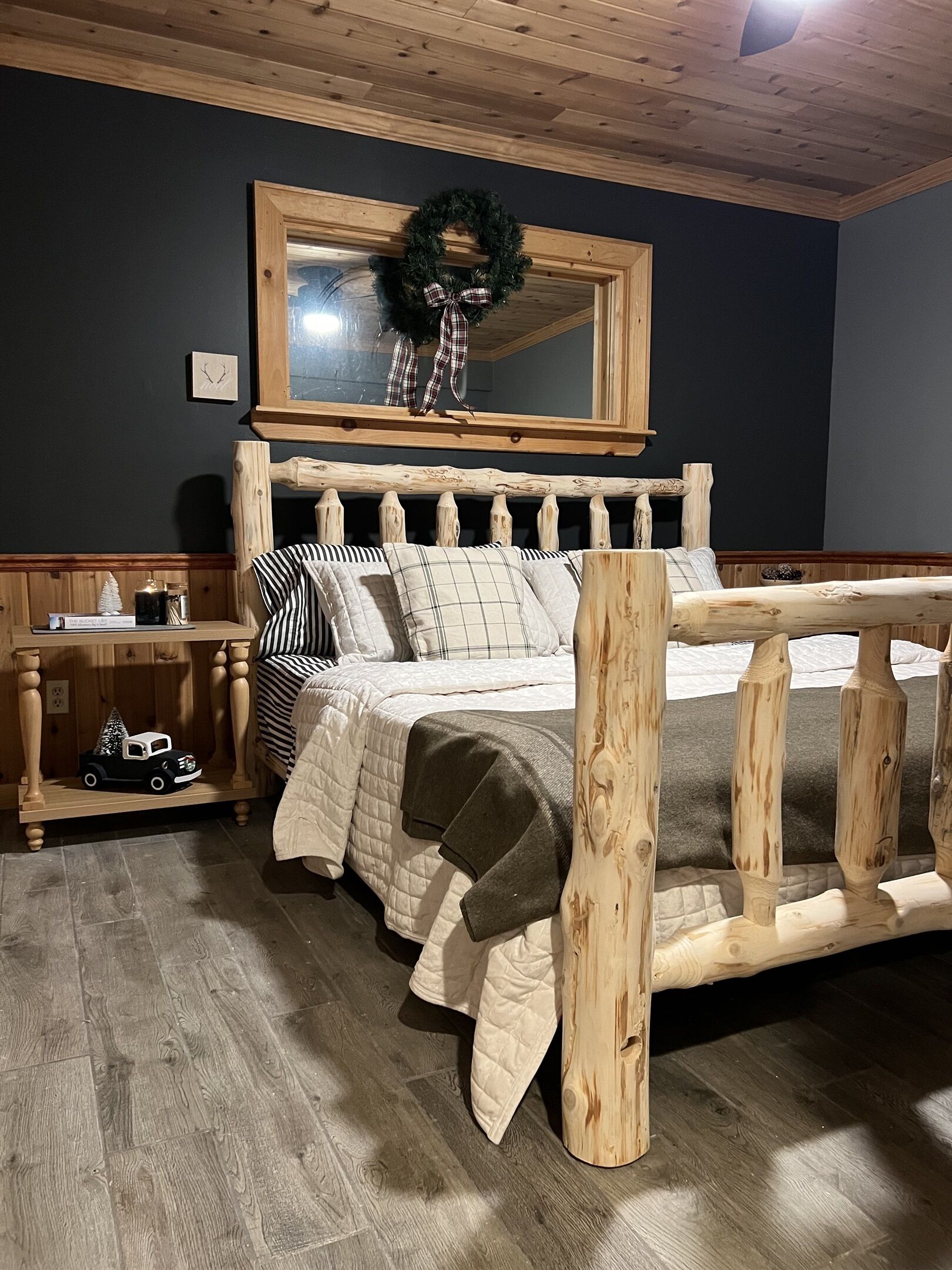 Una cama de madera con cabecera, una mesita de noche con un reloj, y un pequeño estante con un camión de juguete y un árbol de Navidad.