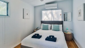 A bedroom with a bed, two pillows, a bedside lamp, a small table, and a wall-mounted air conditioner.