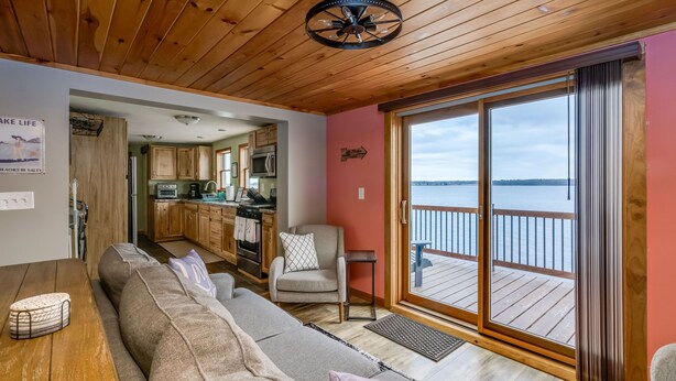 A cozy living room with a wooden ceiling, a sofa, a chair, and a view of a water body through large sliding glass doors. - Black Bay Chalet- Lake front home with magnificent views (Dexter)