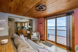 A cozy living room with a wooden ceiling, a sofa, a chair, and a view of a water body through large sliding glass doors. - Black Bay Chalet- Lake front home with magnificent views (Dexter)