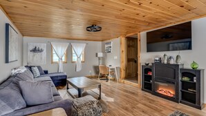 A cozy living room with a wooden ceiling, a grey sofa, a coffee table, a fireplace, and a TV mounted on the wall. - Black Bay Chalet- Lake front home with magnificent views (Dexter)