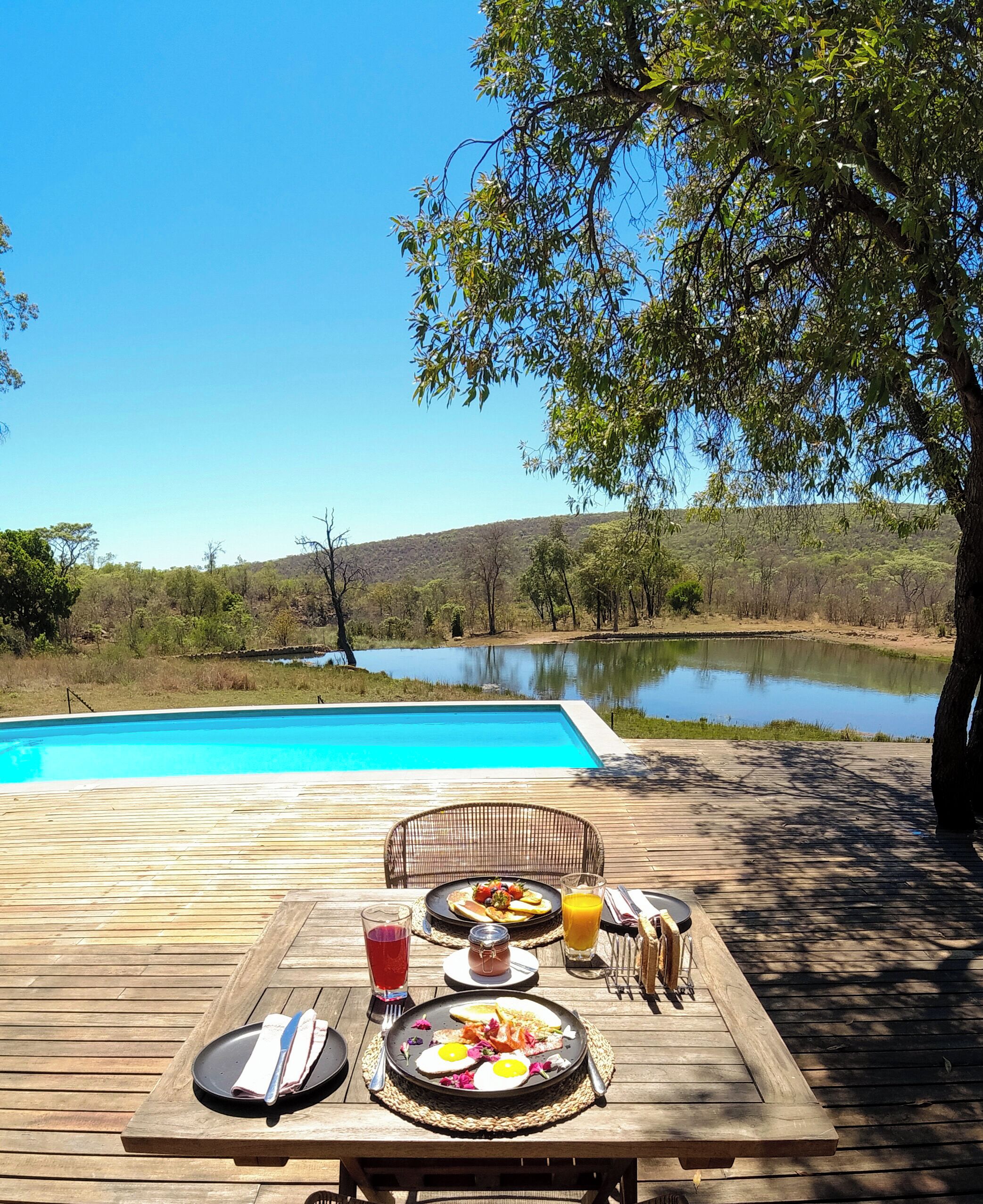 A wooden deck with a table set for breakfast overlooking a swimming pool and a lake.