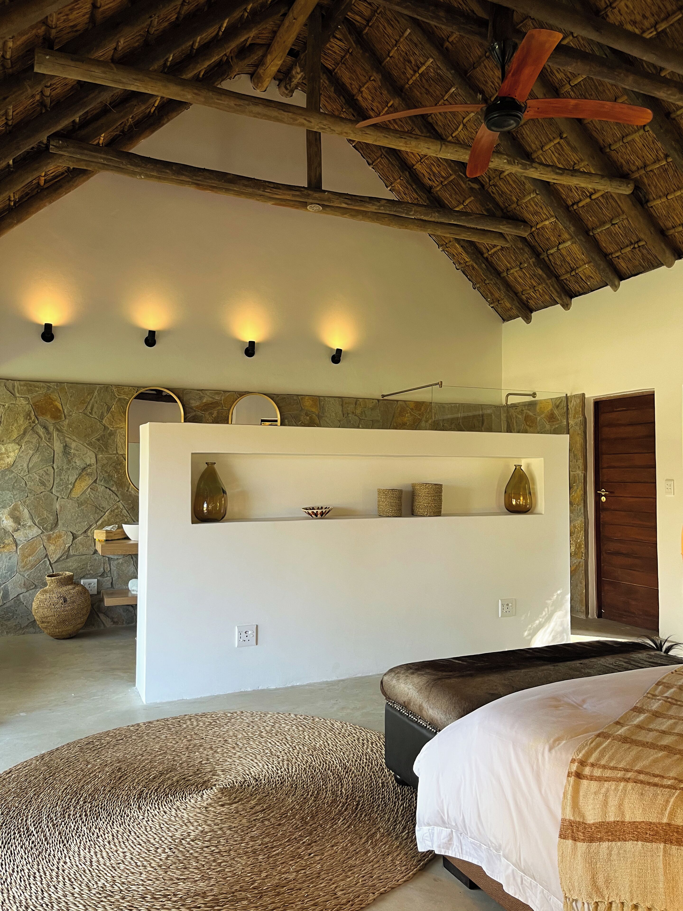 A bedroom with a stone wall, a wooden ceiling, a bed, a bedside table, a mirror, and a shelf with decorative items.