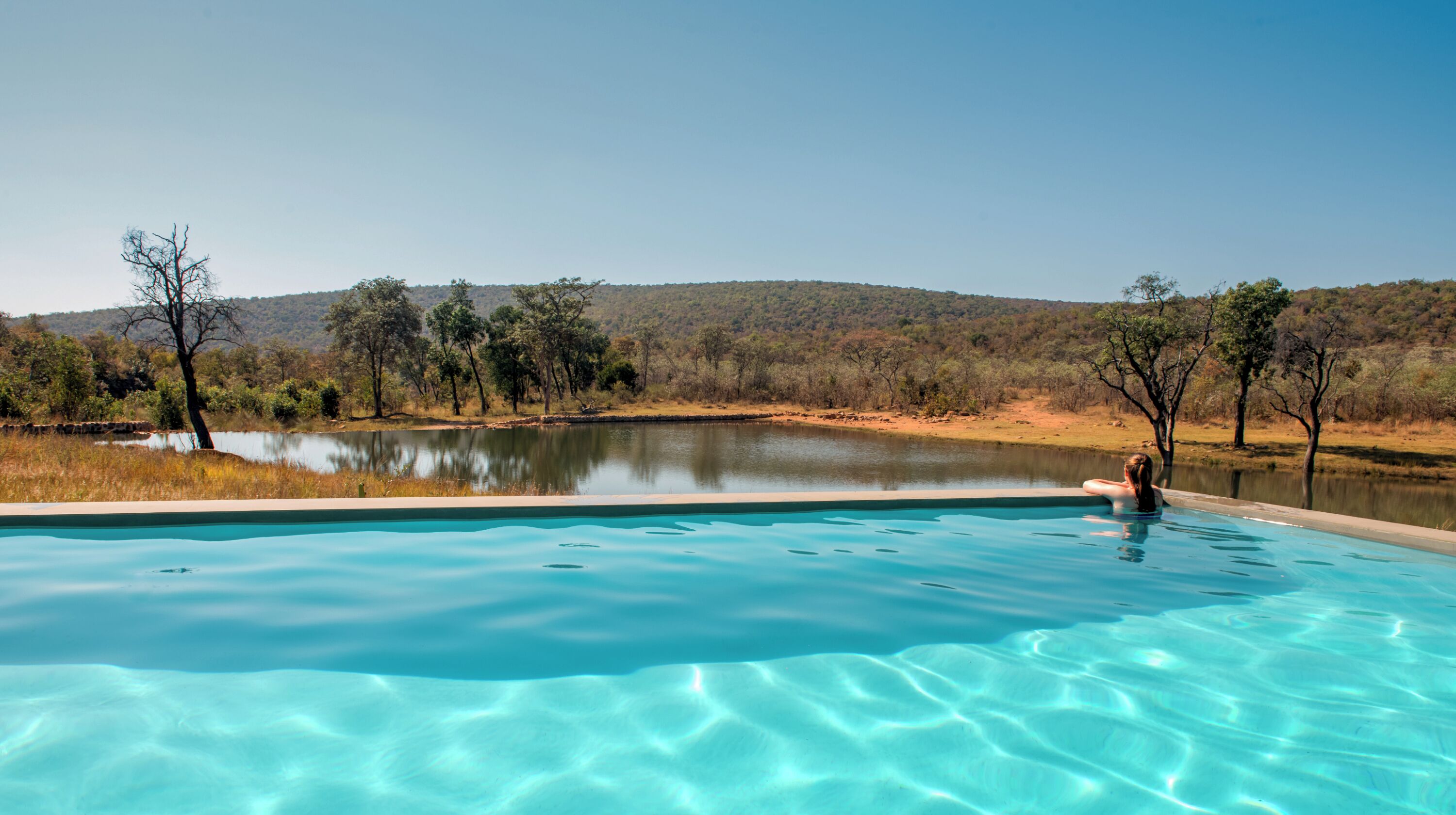 A clear swimming pool with a view of a lake and trees in the background.