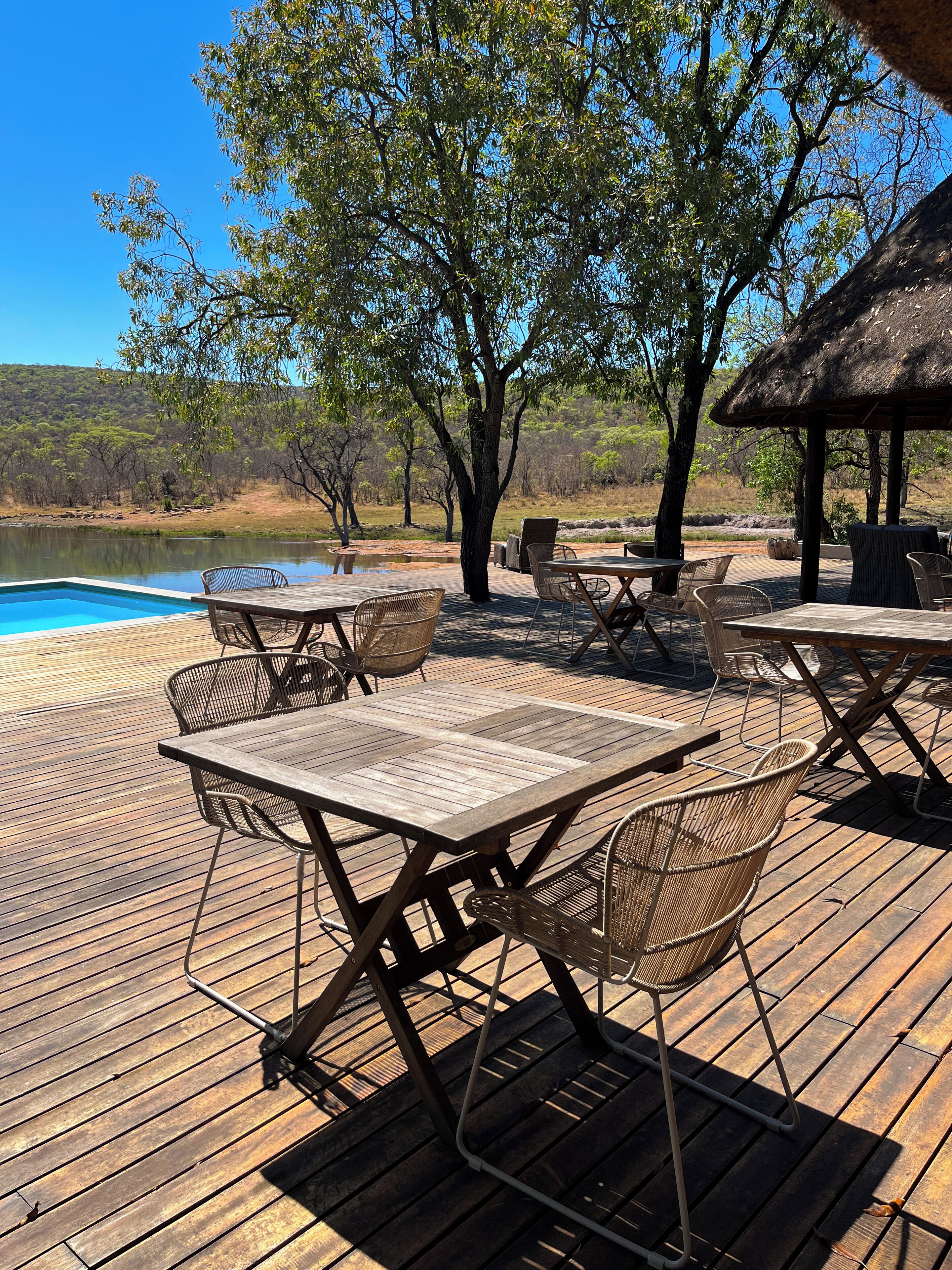 An outdoor deck with a pool, wooden tables, wicker chairs, and a thatched-roof structure.