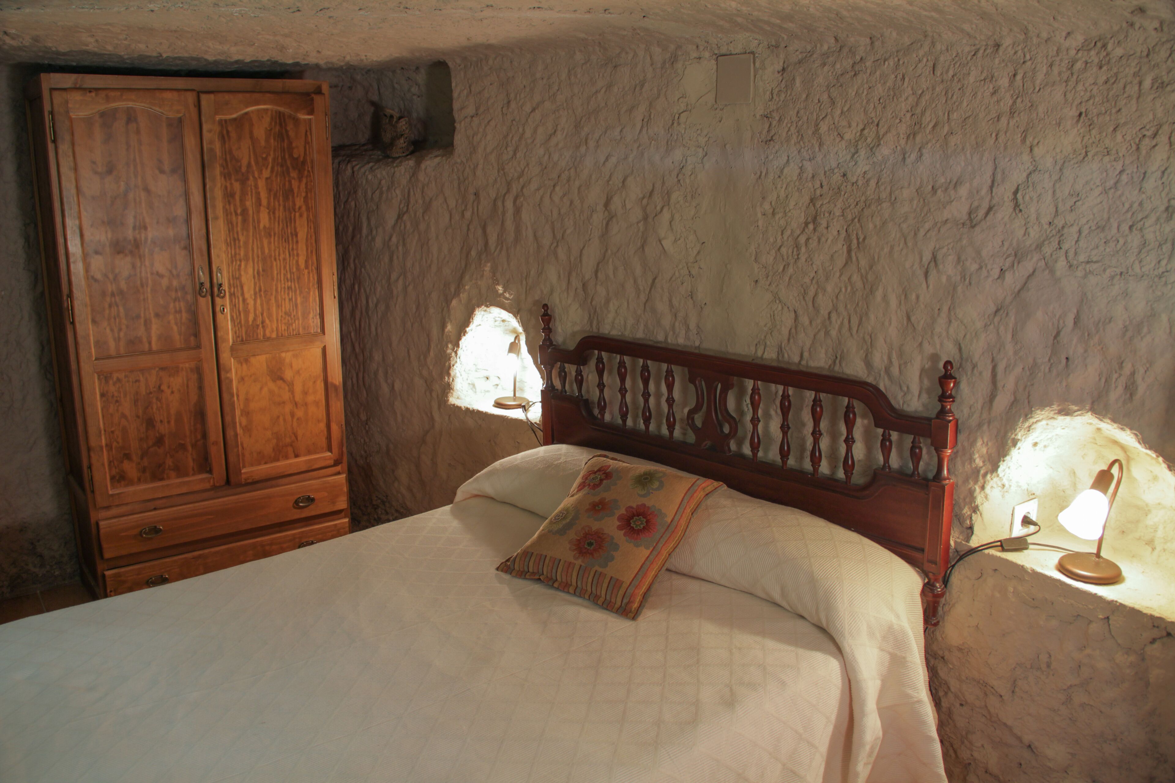 A bedroom with a wooden wardrobe, a bed with a floral pillow, and a wall-mounted lamp.
