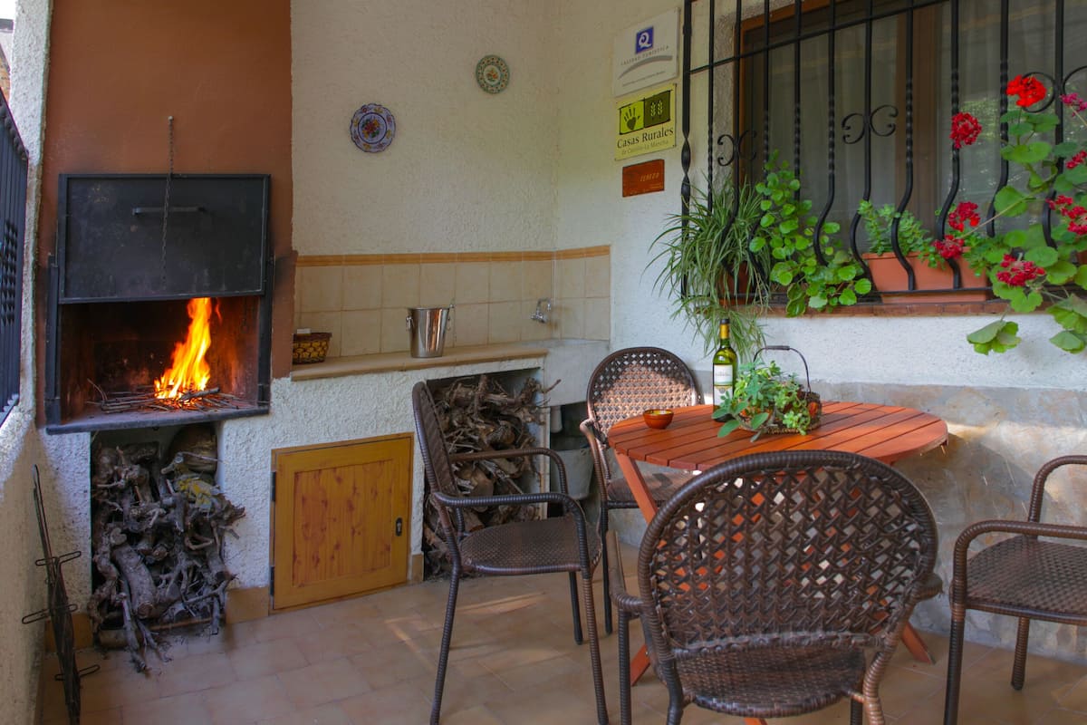 Un patio con chimenea, sillas de mimbre, una mesa de madera y plantas en macetas.