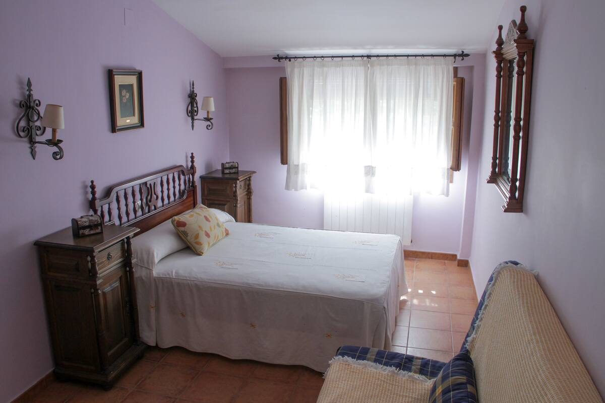 Un dormitorio con cama, mesita de noche, cómoda, ventana con cortinas y espejo en la pared.