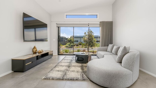 A modern living room with a large flat-screen TV, a grey sofa, a coffee table, and a view of a building and trees through large windows.