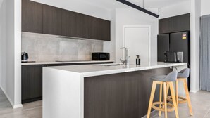 A modern kitchen with dark cabinetry, a white countertop, a built-in microwave, a coffee machine, and a refrigerator. There are two wooden stools at the island.