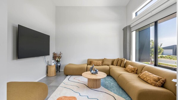 Un salon moderne avec un grand canapé d’angle, une table basse ronde et un téléviseur à écran plat fixé au mur.