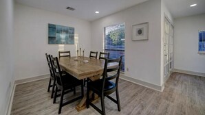 Un coin repas avec une table et des chaises en bois, un tableau au mur et une fenêtre avec des stores.